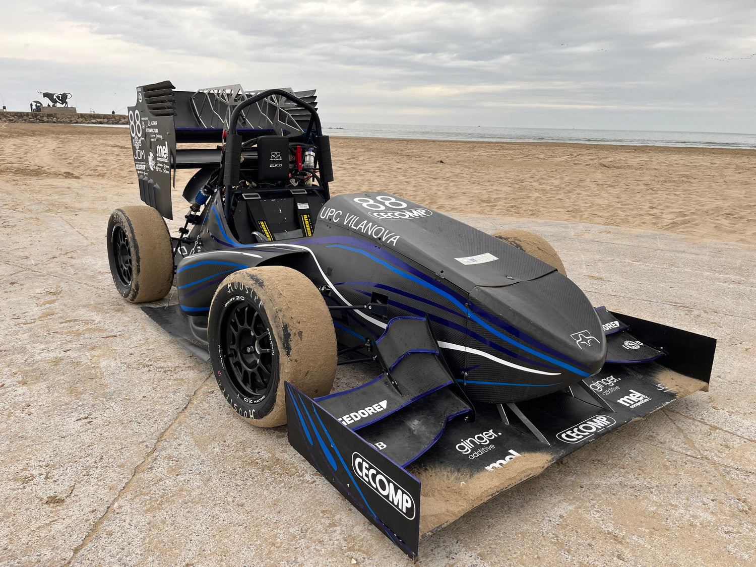 A black and blue formula-style student race car parked on a sandy beach, with sand covering its tires and body panels.